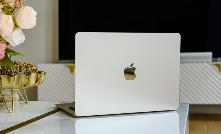 Ein silbernes MacBook mit einem Apple-Logo auf der Rückseite, präsentiert auf einem Glastisch in einem stilvoll dekorierten Raum, neben einer goldenen Dekoschale und weißen Blumen.