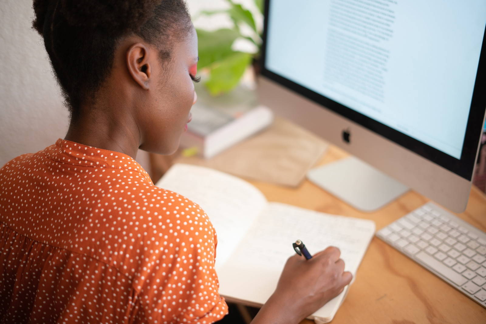 A Woman at a desk in front of her computer, writing in a notebook