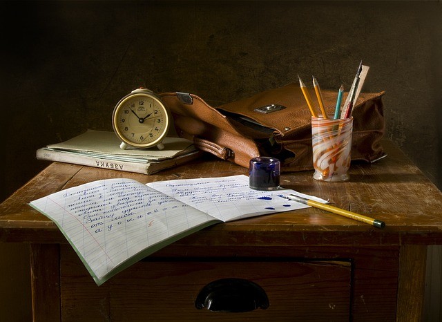 A desk with books, satchel, pens and pencils. There is a notebook opened and a pen next to a split inkwell. Vaia Magazine
